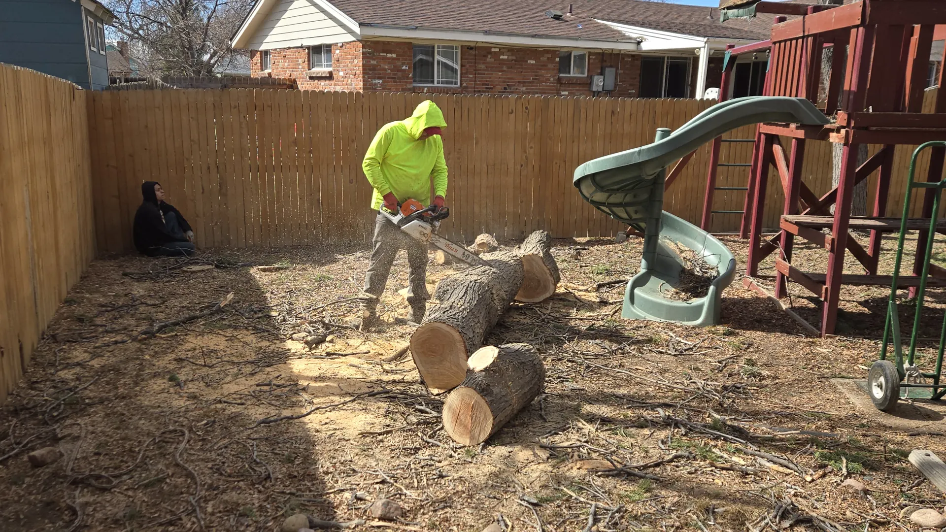 Arbol De Fuego crew member bucking a felled trunk into rounds in a fenced backyard beside a kids' playset